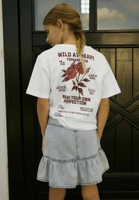 Balta T-kreklu ar sarkaniem grafikām un tekstu aizmugurē, kombinēta ar gaišu džinsu svārku, kura apakšdaļa ir ar viļņotu malu.