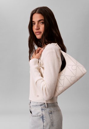 Žena s dlhými tmavými vlasmi v krémovom rebrovanom svetri a svetlých denimových džínsach, nesúca bielu perforovanú kabelku cez rameno.