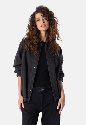 Kvinde med langt krøllet hår iført en sort denimjakke og sorte bukser, stående mod en ensfarvet lys baggrund.