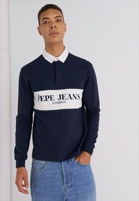Polo de manga larga de color navy con una banda blanca en el pecho que presenta el texto "PEPE JEANS LONDON" y un cuello abotonado, combinado con unos jeans de color azul claro.