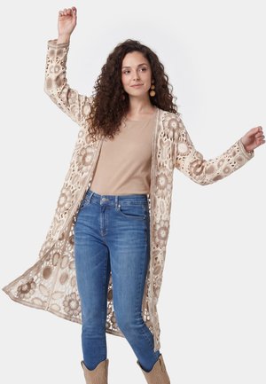 Kvinde med krøllet hår iført en blomsterbroderet cardigan, beige top, blå jeans og brune støvler, løfter armene og ser til siden.