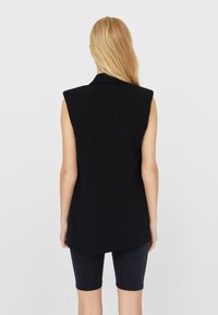 Black sleeveless vest with a straight cut and sharp shoulders, featuring a collar and a smooth texture. Worn over black shorts.