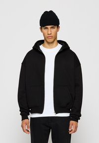 Haut à capuche noir zippé avec une grande capuche, poches avant et poignets côtelés. Porté sur un t-shirt blanc uni, associé à un jean noir.
