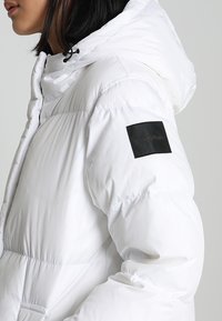 Giacca a vento bianca con design trapuntato, colletto alto, cappuccio regolabile e una patch in tessuto nera con logo sulla manica sinistra.