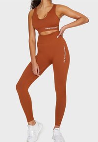 Brązowy zestaw odzieży sportowej z prążkowanej tkaniny, składający się z krótkiego topu i legginsów z wysokim stanem, z białym logo "SQUATPROOF" na pasku.