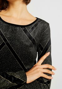 Schwarzes Samtoberteil mit goldenen geometrischen Mustern und Akzenten, langen Ärmeln und rundem Halsausschnitt. Glatte Textur mit schimmernden Details.