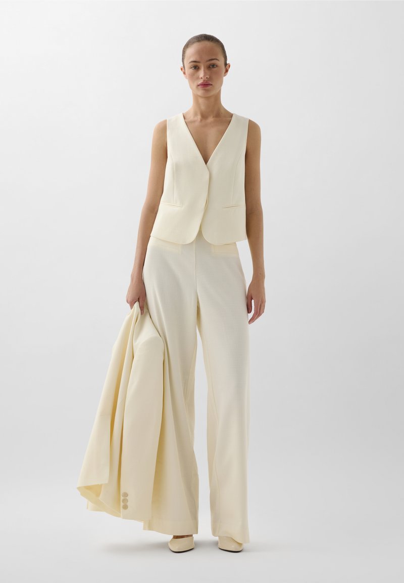 Costume trois pièces jaune pâle avec un gilet ajusté sans manches, un pantalon large et une veste assortie tenue d'une main. Tissu de texture lisse.