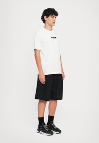 Hvit t-skjorte med korte ermer og en svart etikett på brystet, kombinert med svarte shorts til kneet og svarte joggesko med hvite detaljer.