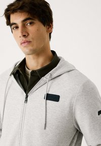 Sudadera gris con cremallera y cordones ajustables, que presenta una tela texturizada, un parche de logo en el pecho y una camiseta oscura en capas debajo.