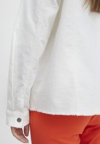 Hvid, tekstureret jakke med en frynset kant og knappet manchet detaljer, parret med klare røde bukser. Nærbillede af ærmet og kanten.