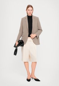 Beige blazer, svart genser med høy hals, lyse kremfargede plissert shorts og svart håndveske. Svart spisseskotnæss fullfører antrekket. Enkelt og moderne design.