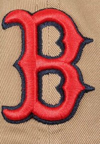 Tan fabric cap featuring a prominent red "B" logo, outlined in dark blue embroidery, with a structured design and textured surface.