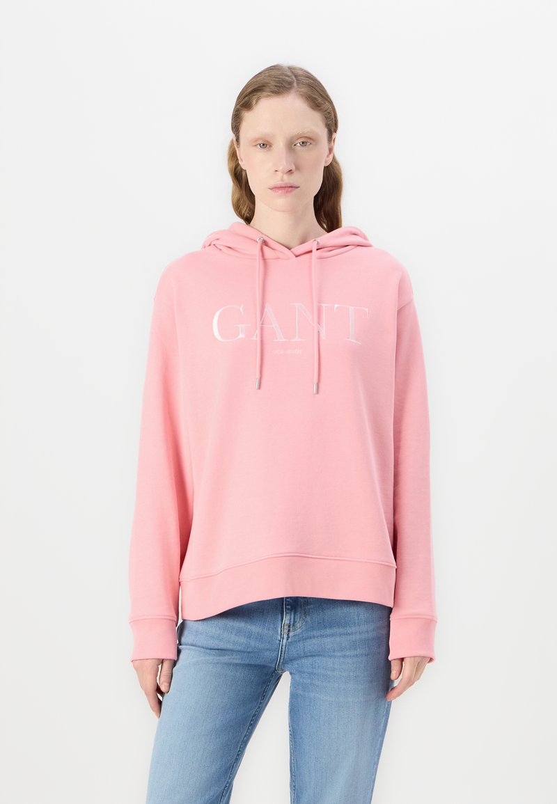 Ljusrosa hoodies med en framstående känguruficka, snörning i huvan och vit "GANT"-logotyp. Mjuk tyg, avslappnad passform och ribbade cuffs.