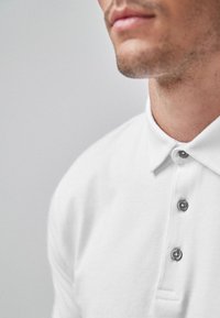 Hvid polo-shirt med klassisk krave og tre-knaps plade, med en glat tekstur og diskret syningsdetalje ved kanten.