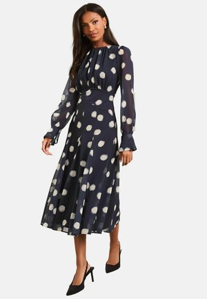 Frau trägt ein nachtblaues Midi-Kleid mit langen transparenten Ärmeln und weißen Punkten, kombiniert mit schwarzen spitz zulaufenden Absatzschuhen.