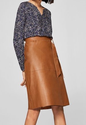 Femme portant une jupe en cuir marron taille haute avec une ceinture nouée et un chemisier bleu marine à fleurs à manches longues.