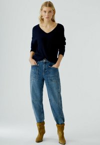 Femme aux cheveux blonds portant un pull noir en V, un jean taille haute bleu avec de grandes poches avant, et des bottines en daim marron, debout.