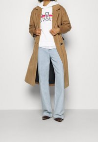 Cappotto di lana cammello con un design strutturato, indossato sopra un felpa bianca con il testo "GAP" multicolore, abbinato a jeans azzurri chiari e stivali marroni.
