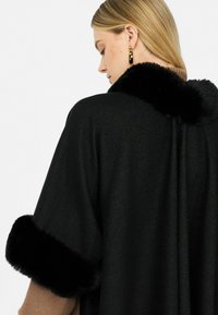 Donna con capelli biondi che indossa uno scialle nero con rifiniture in finta pelliccia sul collo e sulle maniche, abbinato a orecchini a cerchio dorati.