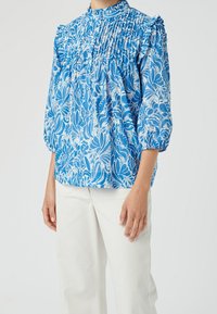 Blouse à imprimé floral bleu avec épaules froncées et col haut, ornée de motifs blancs. Confectionnée en tissu léger, avec des manches bouffantes.