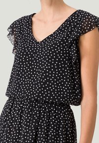 Blusa nera con pois, scollo a V e maniche a volant in tessuto trasparente. Presenta una vita plissettata per un aspetto aderente e un tessuto leggero e strutturato.