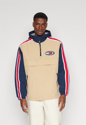 Chaqueta ligera beige con cuello con cremallera, mangas azul marino con rayas rojas y blancas, y un parche de logo en el pecho.