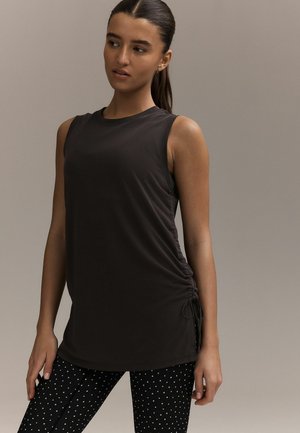 Žena s tmavým copom, oblečená v čiernej bezrukávovej tunike s detailom viazania na boku a čiernych legínach s bielymi bodkami, stojaca na jednoduchom pozadí.