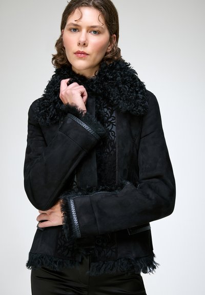 Jeune femme portant une veste noire doublée de fausse fourrure, touchant le col d'une main, expression faciale neutre, fond clair.