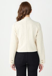 Cremefarbene Tweedjacke mit kurzem Schnitt, langen Ärmeln und drei goldenen Knöpfen an den Manschetten. Schlichtes Rückendesign.