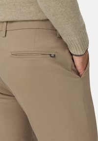 Pantalones beige con una textura suave, que cuentan con un bolsillo con cremallera en la parte trasera y un corte ajustado, con detalles de costura y un acento de logo.