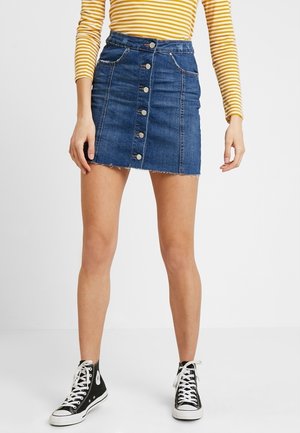 Henkilö pukeutuneena keltaiseen raidalliseen pitkähihaseen paitaan, siniseen denim-napilliseen minihameeseen ja mustiin korkeavartisiin tennareihin, seisoo sisätiloissa.