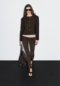 Vrouw in een donkergrote jas, suède broeken, sandalen en zonnebril, met een grote leren schoudertas voor een effen achtergrond.