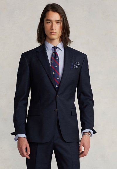 Mørk marineblå dressjakke med to knapper, hakkede slag og matchende bukser. Lyseblå skjorte og mønstret slips med aksenter. Lommetørkle inkludert.