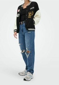 Czarna i biała kurtka varsity z naszywkami, czarny top na ramiączkach, zniszczone niebieskie dżinsy z dziurami oraz srebrne buty sportowe.