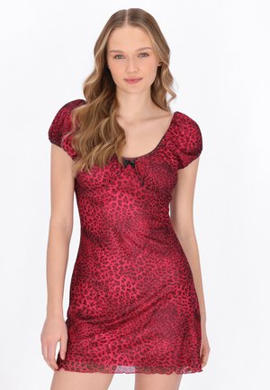 Kvinde med langt, bølget blondt hår, iført en kort kjole med rødt og sort leopardprint og pufærmer, stående mod en hvid baggrund.