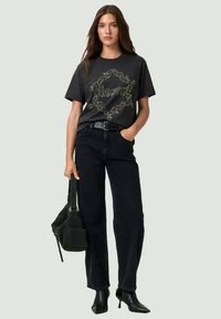 Black graphic t-shirt with ornate gold design, paired with high-waisted black jeans, ankle boots, and a textured shoulder bag.