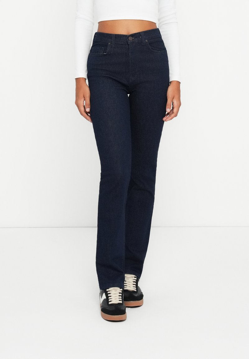 Jeans de tiro alto de color azul oscuro con una silueta ajustada, dos bolsillos frontales y un cierre de cremallera. Combinados con zapatillas negras con cordones blancos.