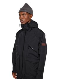 Mann trägt eine schwarze Kapuzenjacke mit mehreren Taschen und eine graue Strickmütze vor weißem Hintergrund.