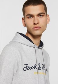 Grå hoodie med snøre og tofarvet stof. Har "Jack & Jones" logo i marineblå og gul broderi, glat tekstur.