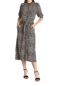 Žena v šatech midi s leopardím potiskem, krátkými nafouklými rukávy, předním zapínáním na knoflíky, páskem v oblasti pasu a černými kotníkovými sandály.