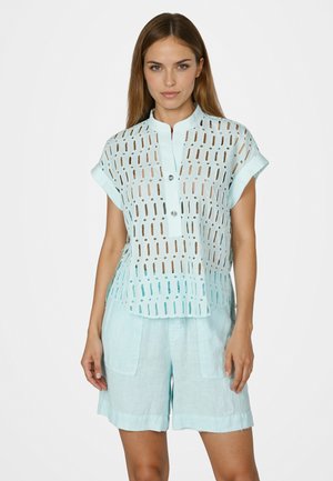 Woman with long hair wearing light blue perforated short-sleeve blouse and matching shorts, standing against a plain white background.