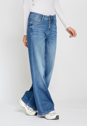 Personne portant un jean large bleu clair, un haut à manches longues blanc et des baskets blanches, debout devant un fond uni.