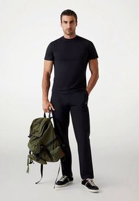 T-shirt de sport noir, style ajusté, associé à un pantalon noir. Tient un sac à dos vert olive avec des sangles noires et une poche latérale. Baskets noires.