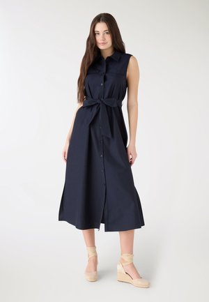 Donna con capelli lunghi castani che indossa un vestito midi blu navy senza maniche, con bottoni e cintura in vita, abbinato a zeppe espadrillas beige, su sfondo bianco.