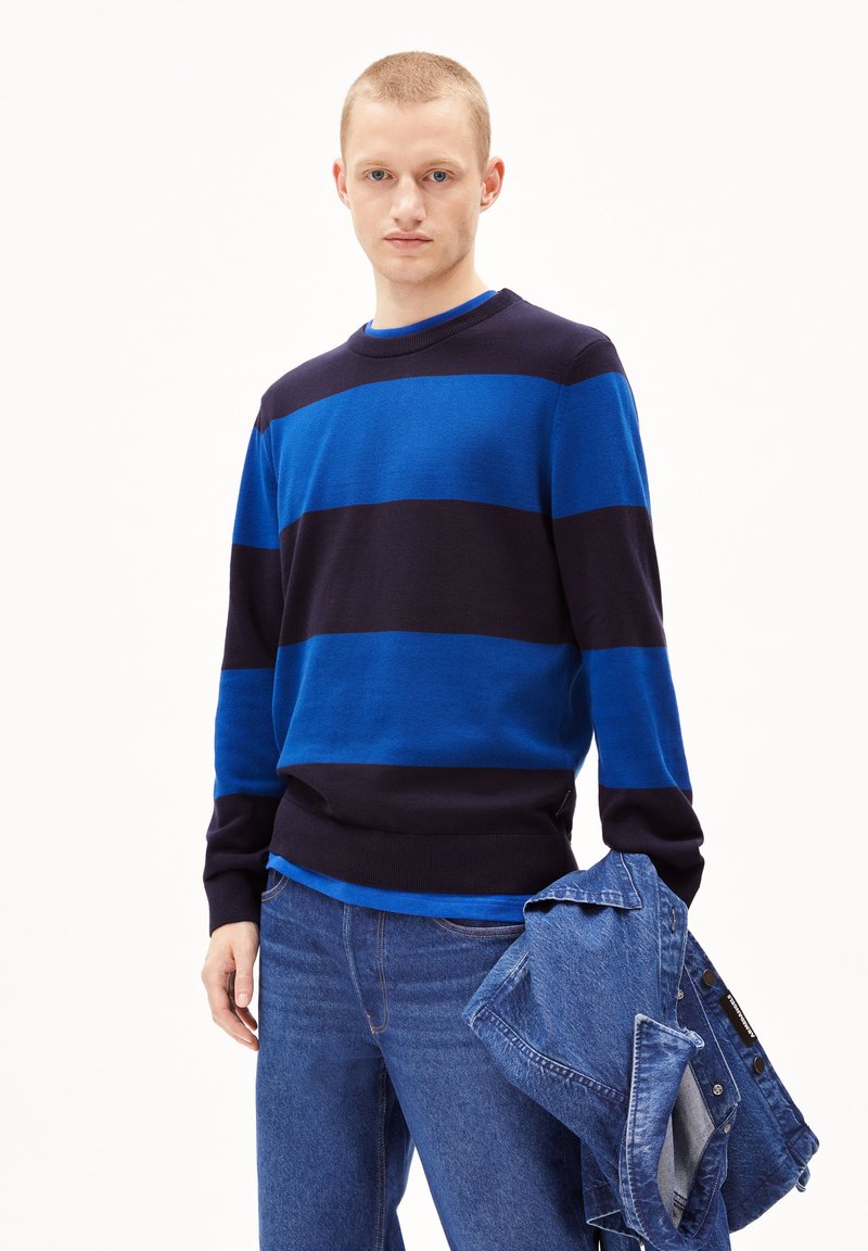Gestreifter Pullover in Blau und Dunkelnavy, Strickstoff, runder Halsausschnitt, lange Ärmel, getragen mit blauen Jeans, hält eine Jeansjacke.