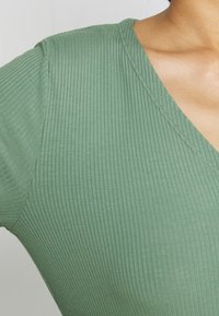 Zielony, prążkowany top z dekoltem w serek, krótkimi rękawami i miękką teksturą. Materiał jest gładki z delikatnymi pionowymi liniami.