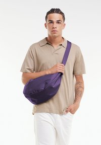 Borsa a tracolla viola con una texture liscia, forma arrotondata e tracolla regolabile. Indossata a traverso sul corpo sopra una maglietta polo beige.