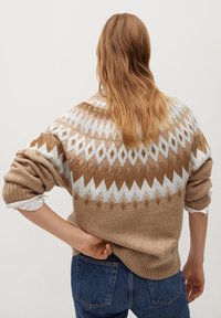 Überdimensionierter cremefarbener Pullover mit braunen geometrischen Mustern. Mit gerippten Bündchen und Saum, weichem Material und lässiger Passform, getragen über blauen Jeans.