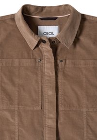 Chemise marron à boutons avec col et deux poches avant, avec une étiquette blanche visible portant le nom de la marque "CECIL".