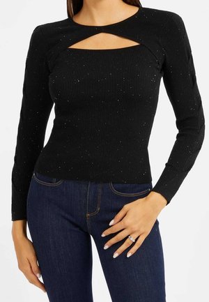 Kvinde iført sort ribbet trøje med lange ærmer, udskæring ved brystet og subtile glimmer, kombineret med mørkeblå jeans, hånden hviler på hoften.